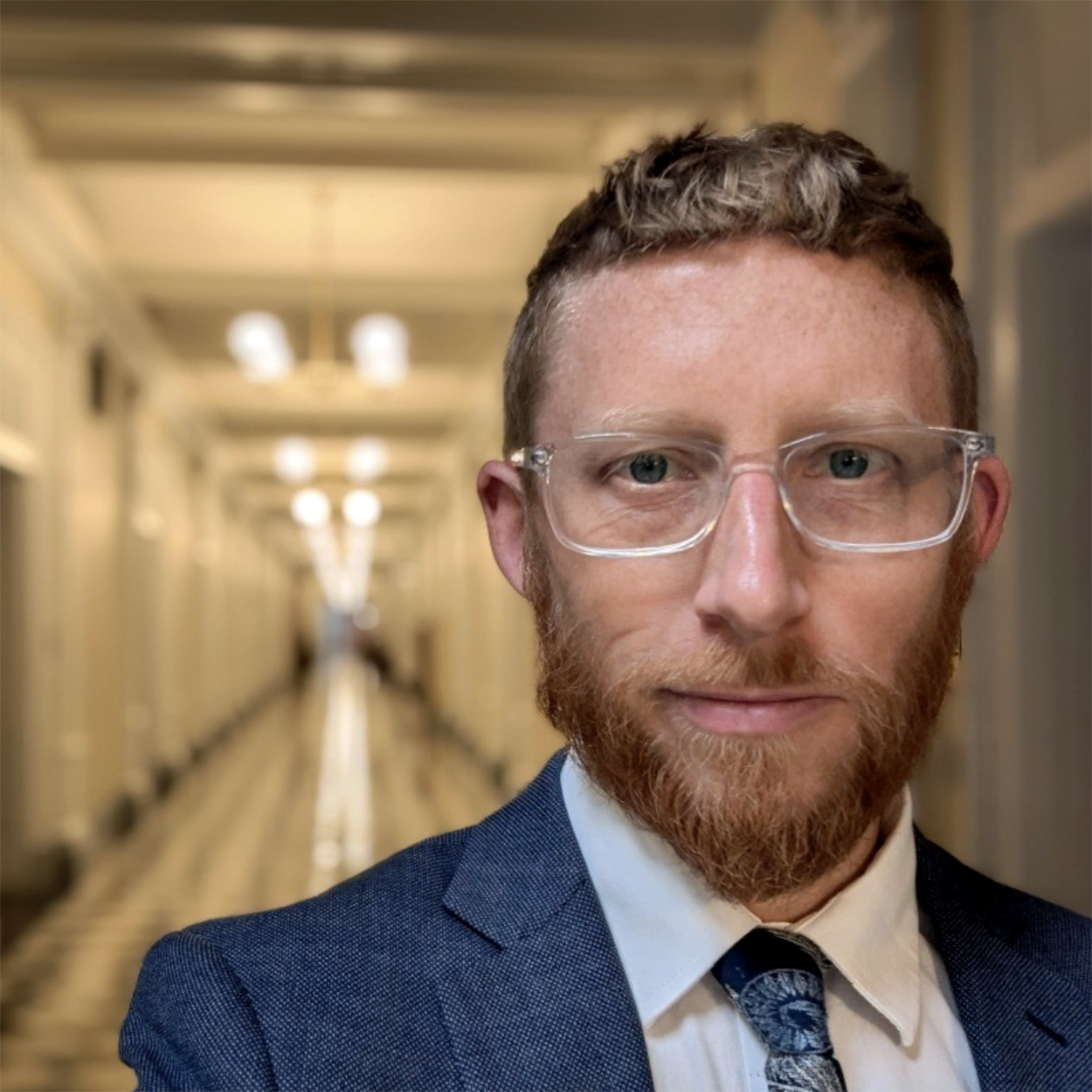 Ben Beachy, Senior Advisor at Public Renewables Project, wearing a blue suit, white shirt, and patterned tie, standing in a well-lit hallway with a blurred background.